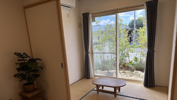 A room with a sliding door leading to a balcony, a tatami mat floor, a small wooden table, and a potted plant.