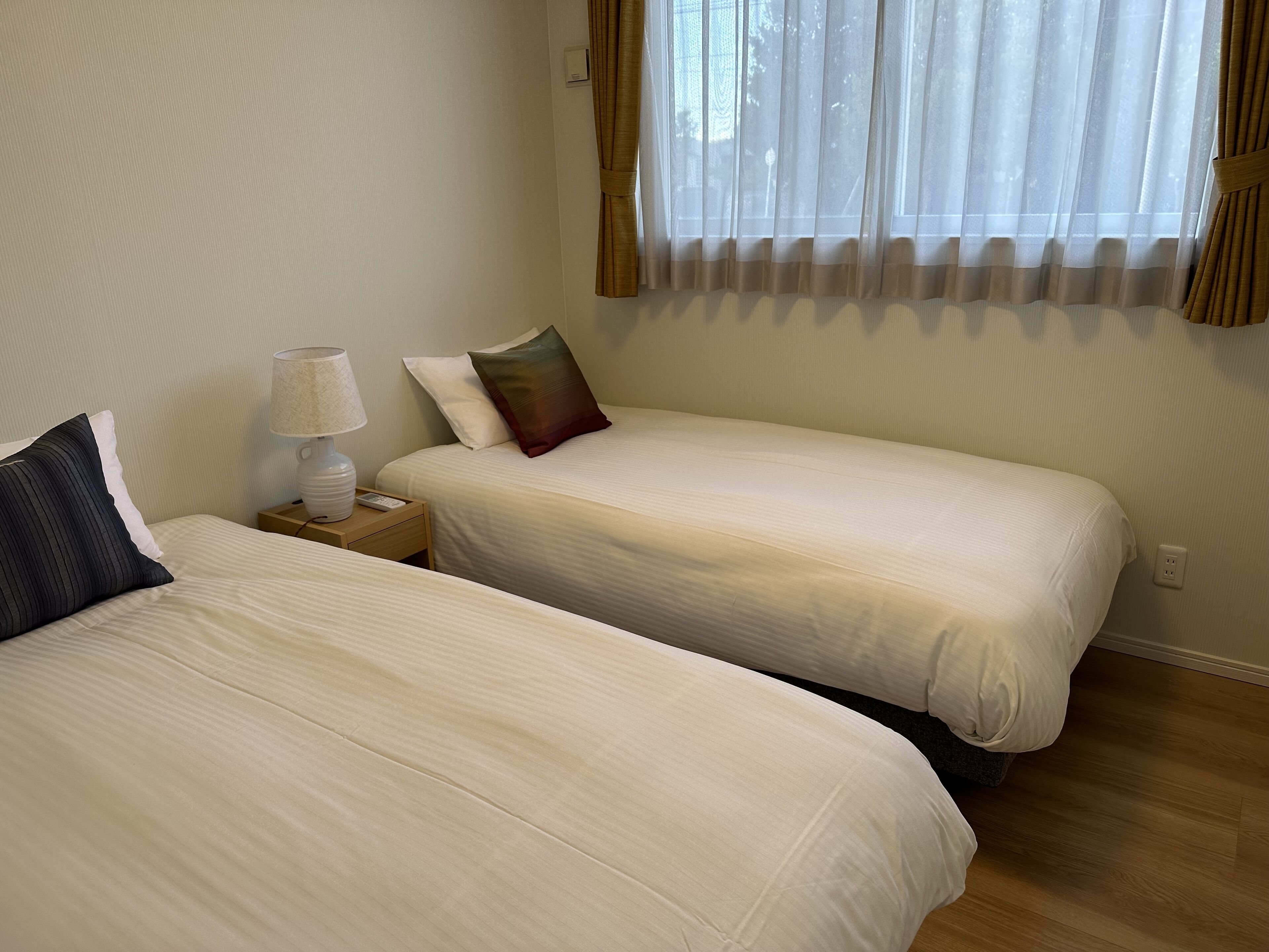 Une chambre à deux lits, avec deux lits simples, une table de chevet avec une lampe et une fenêtre ornée de rideaux légers.