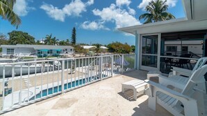 Un balcone con piscina, sedie a sdraio e vista sull'area circostante, tra palme e altri edifici.