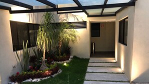 Un área exterior moderna con un sendero de hormigón, un pequeño césped y diversas plantas, incluyendo una palmera.