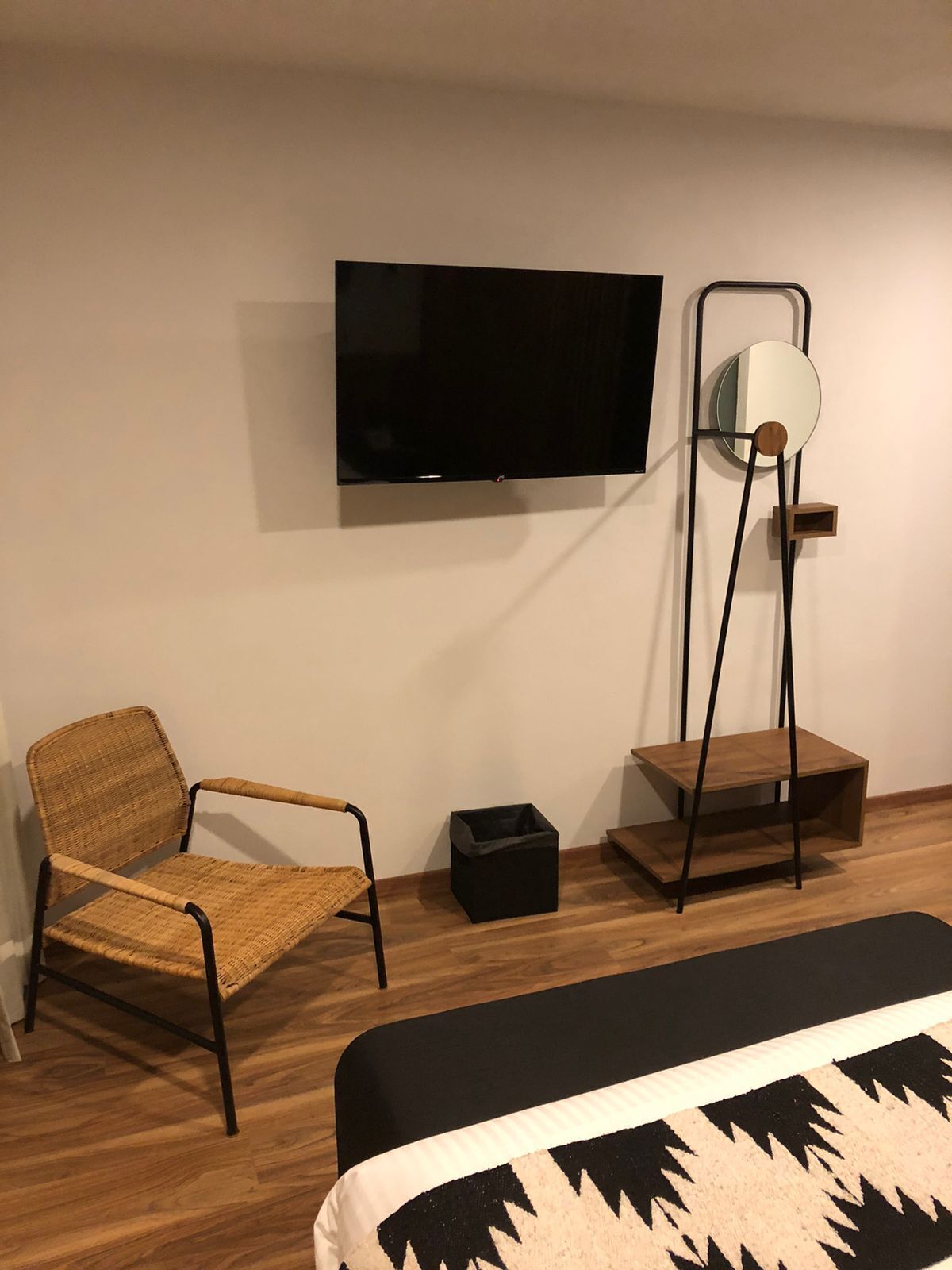 Ein modernes Zimmer mit einem Wand-Fernseher, einem Korbstuhl, einer kleinen schwarzen Mülltonne und einem Nachttisch aus Holz mit Spiegel.