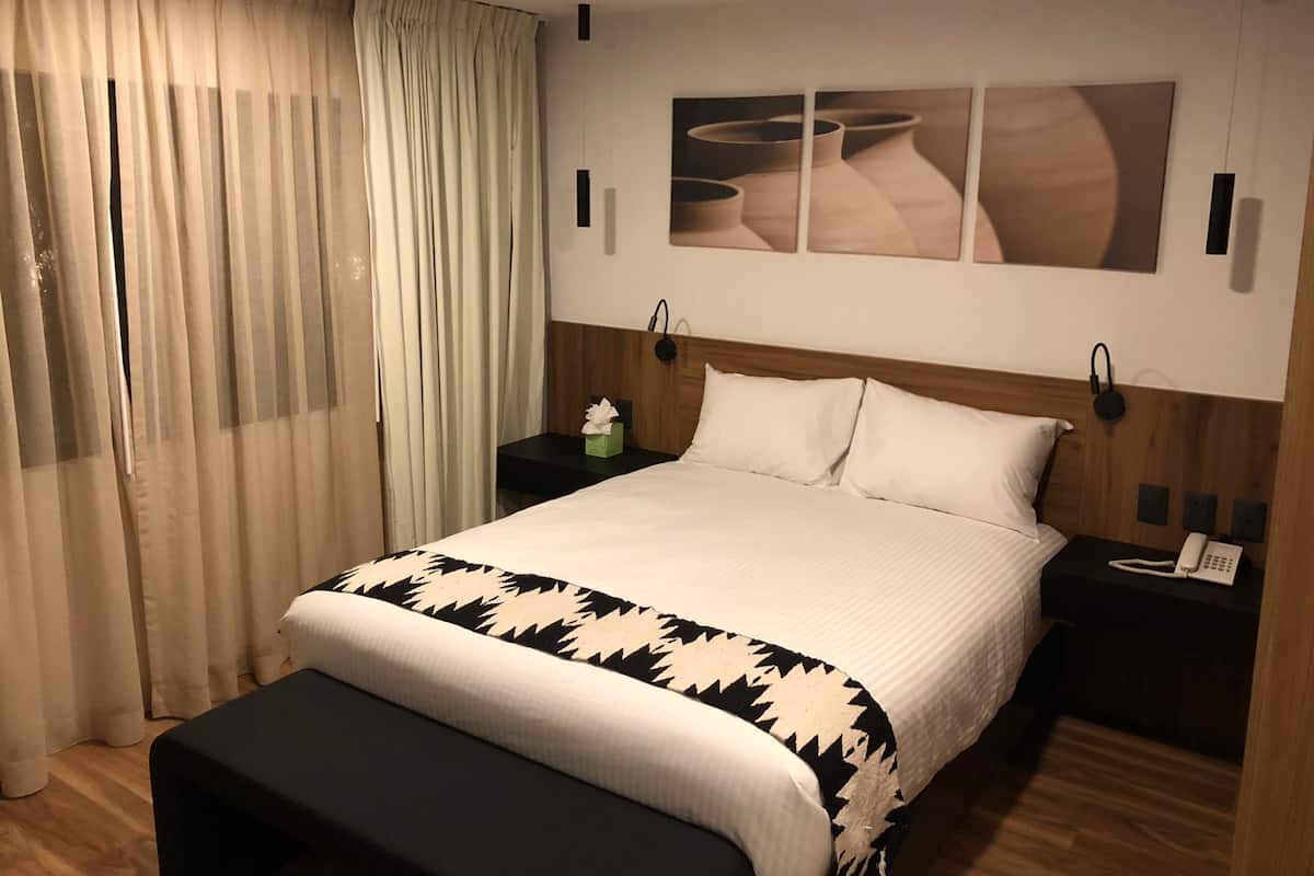 Une chambre d’hôtel avec un grand lit, une table de chevet avec un téléphone, une petite table de chevet avec une boîte de mouchoirs, et une œuvre d’art accrochée au mur au-dessus du lit.