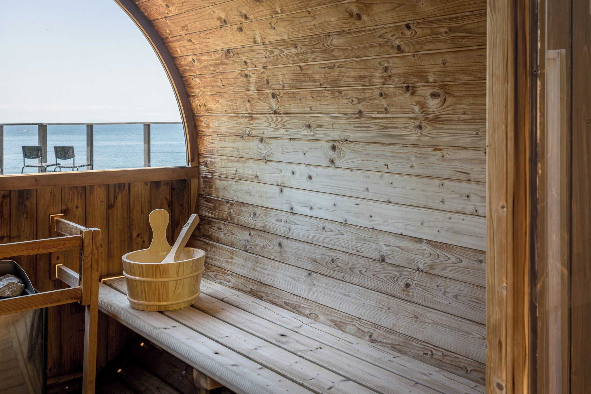 A wooden cabin with a bench and a wooden tub, overlooking a body of water.