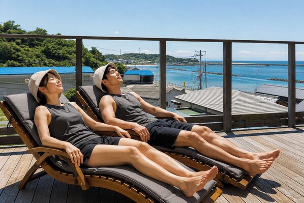 Two people sunbathing on lounge chairs with a view of the sea and a wooden deck.