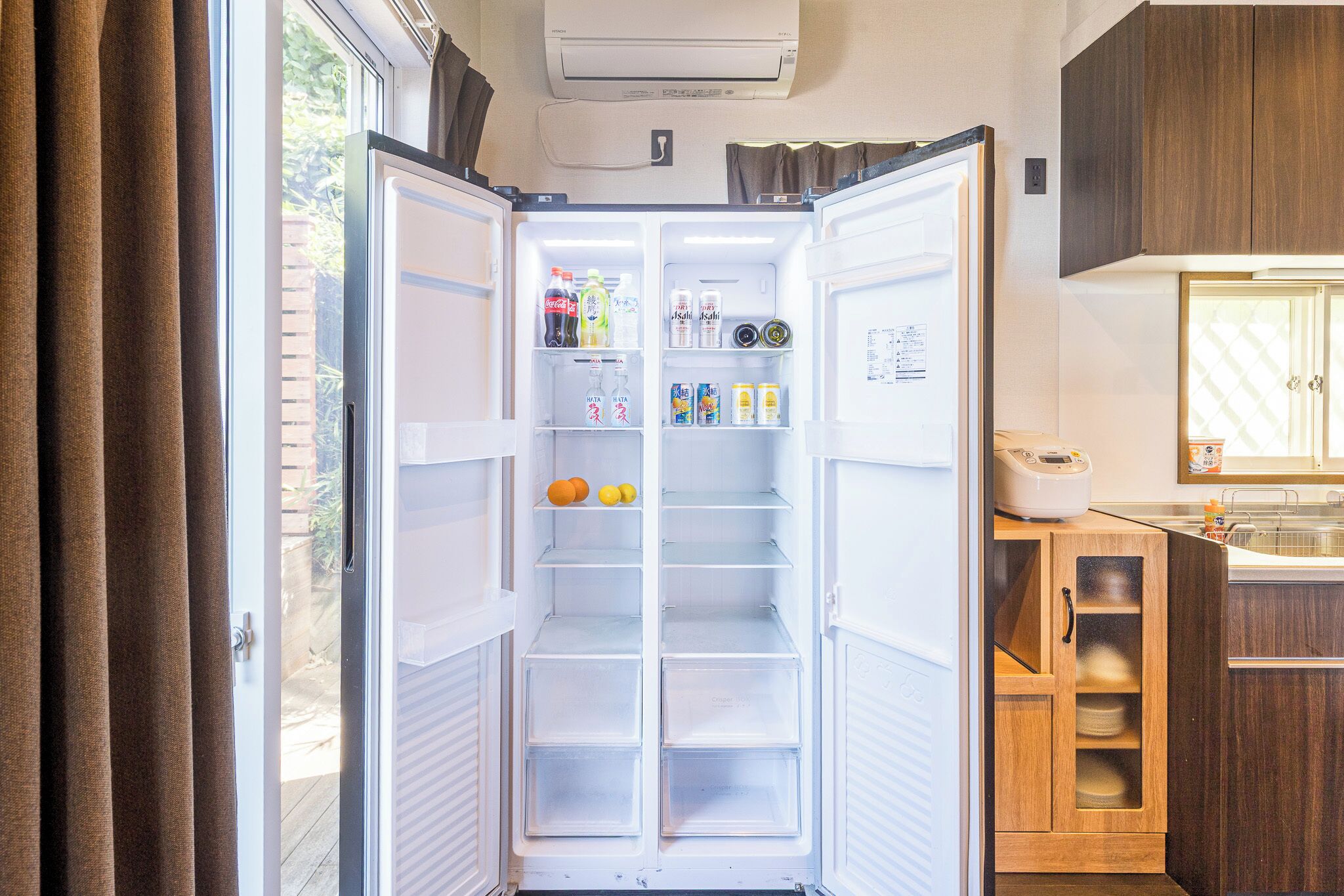 Un réfrigérateur ouvert, rempli de boissons et de fruits, une fenêtre avec des rideaux et un coin cuisine avec des meubles en bois.