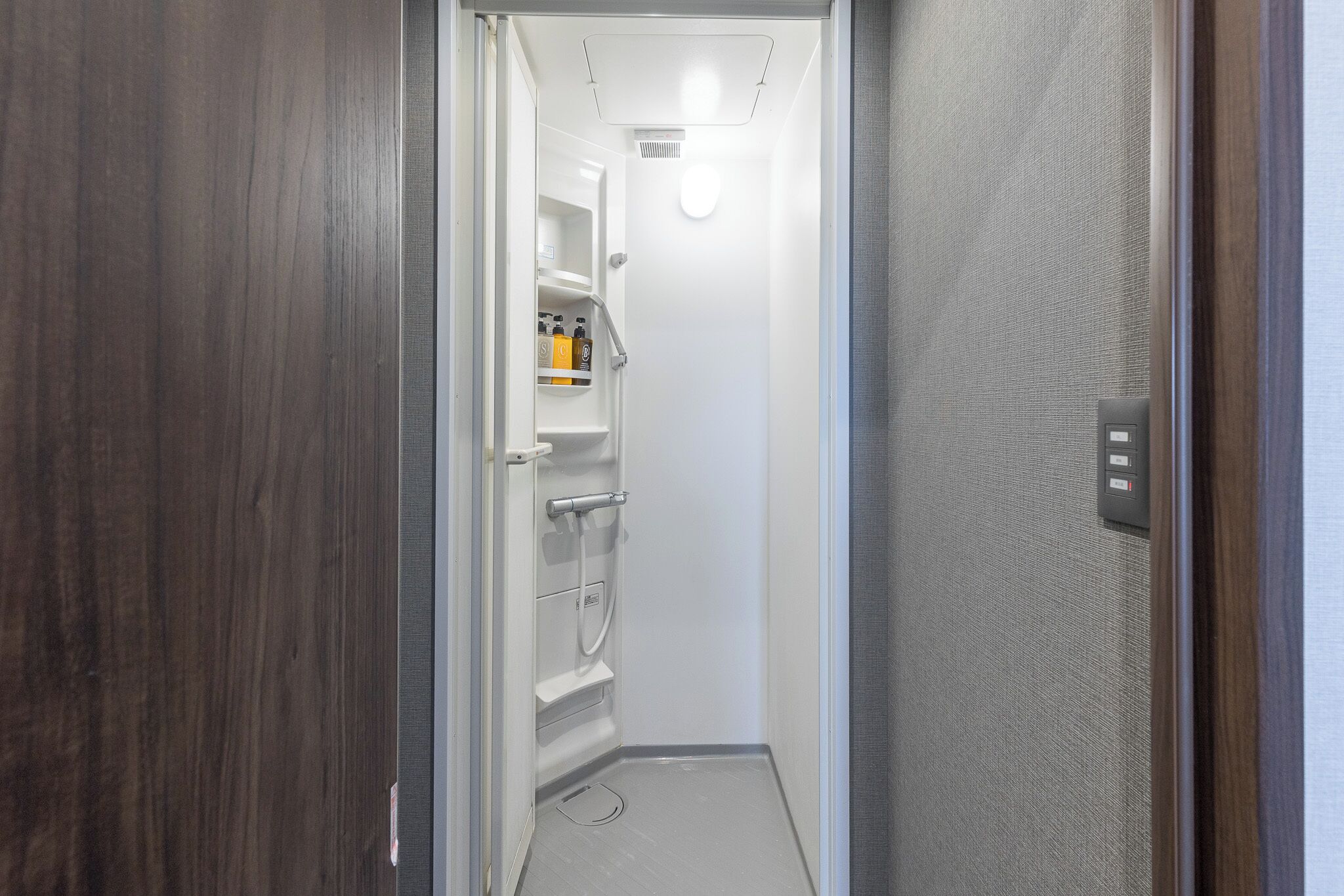 A narrow hallway with a door leading to a room with a white wall, a shelf with toiletries, and a light fixture.