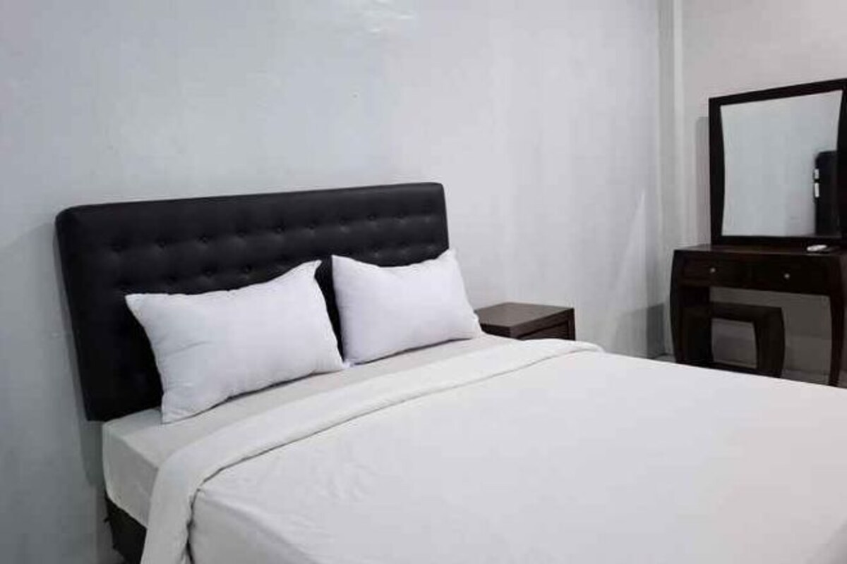 A bed with a black headboard and white bedding in a room with a wooden nightstand and a mirror on a dresser.