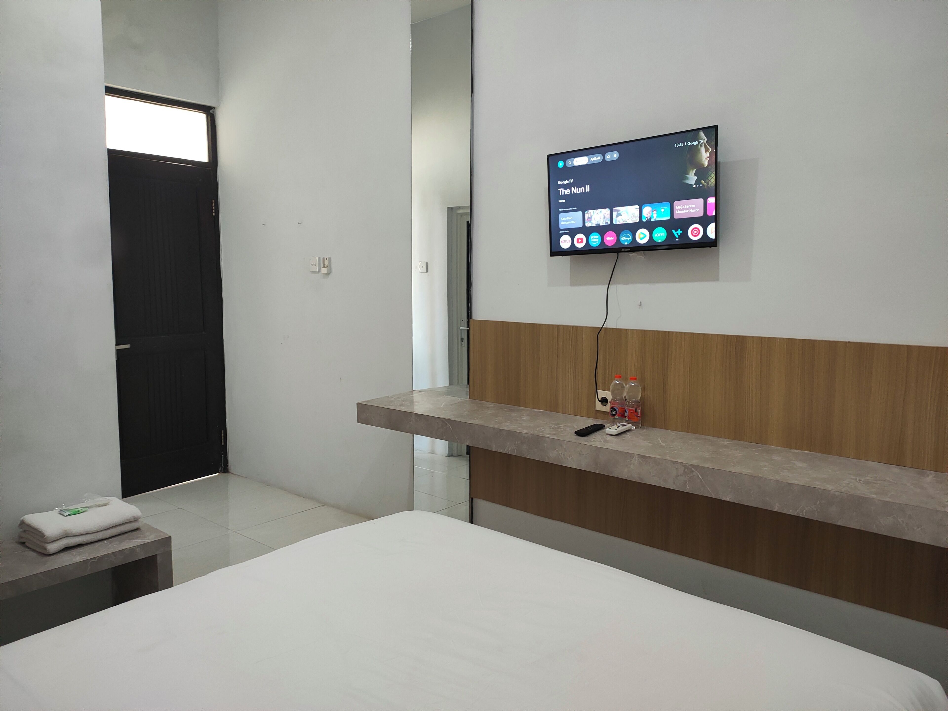 A hotel room with a bed, a bedside table with a remote control, a TV mounted on the wall displaying a streaming service interface, and a wooden panel on the wall.