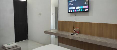 A hotel room with a bed, a bedside table with a remote control, a TV mounted on the wall displaying a streaming service interface, and a wooden panel on the wall.