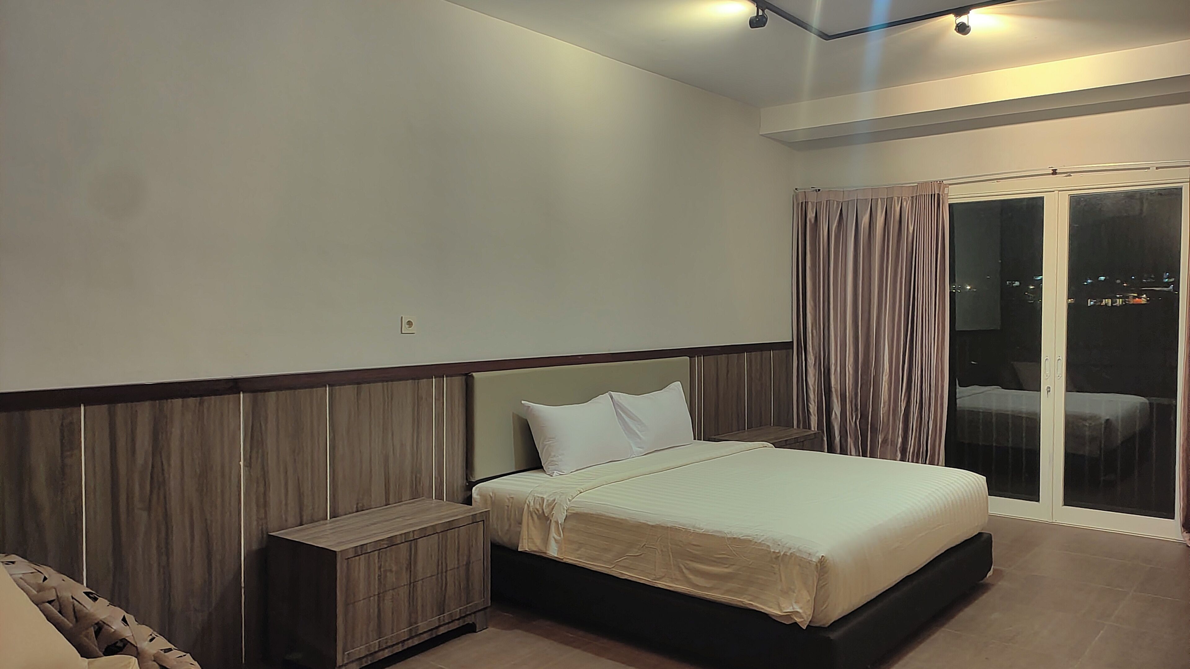 Un dormitorio con cama, mesita de noche, cabecera de madera, puerta de cristal corrediza y vista de la ciudad por la noche.