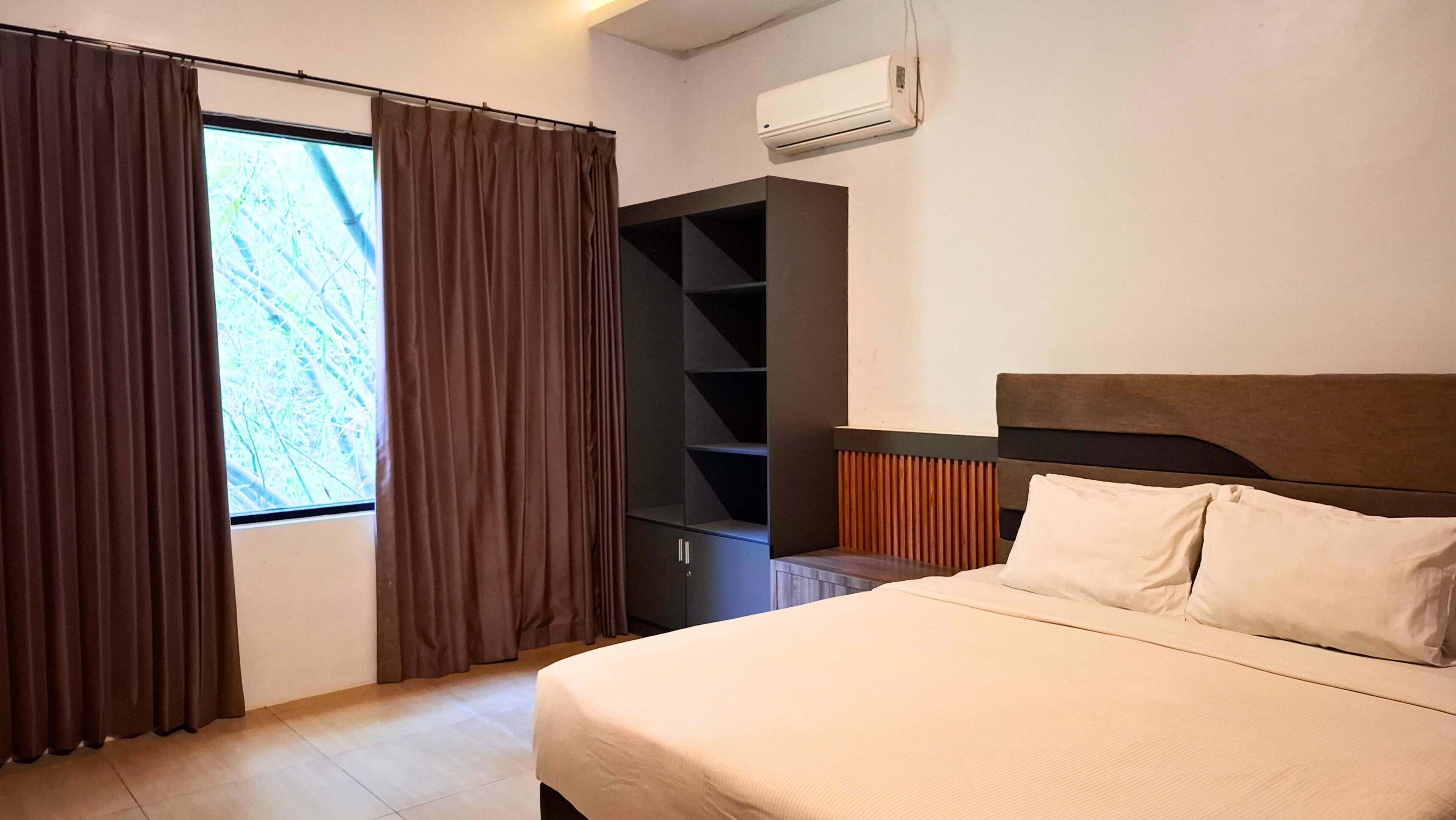 A hotel room with a bed, a wardrobe, a window with curtains, and an air conditioning unit.