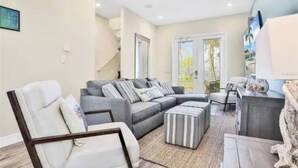 Un salón espacioso con un sofá seccional gris, un sillón blanco y un reposapiés a rayas. Una mesa de centro de madera está en el centro, y un ventanal grande proporciona luz natural.