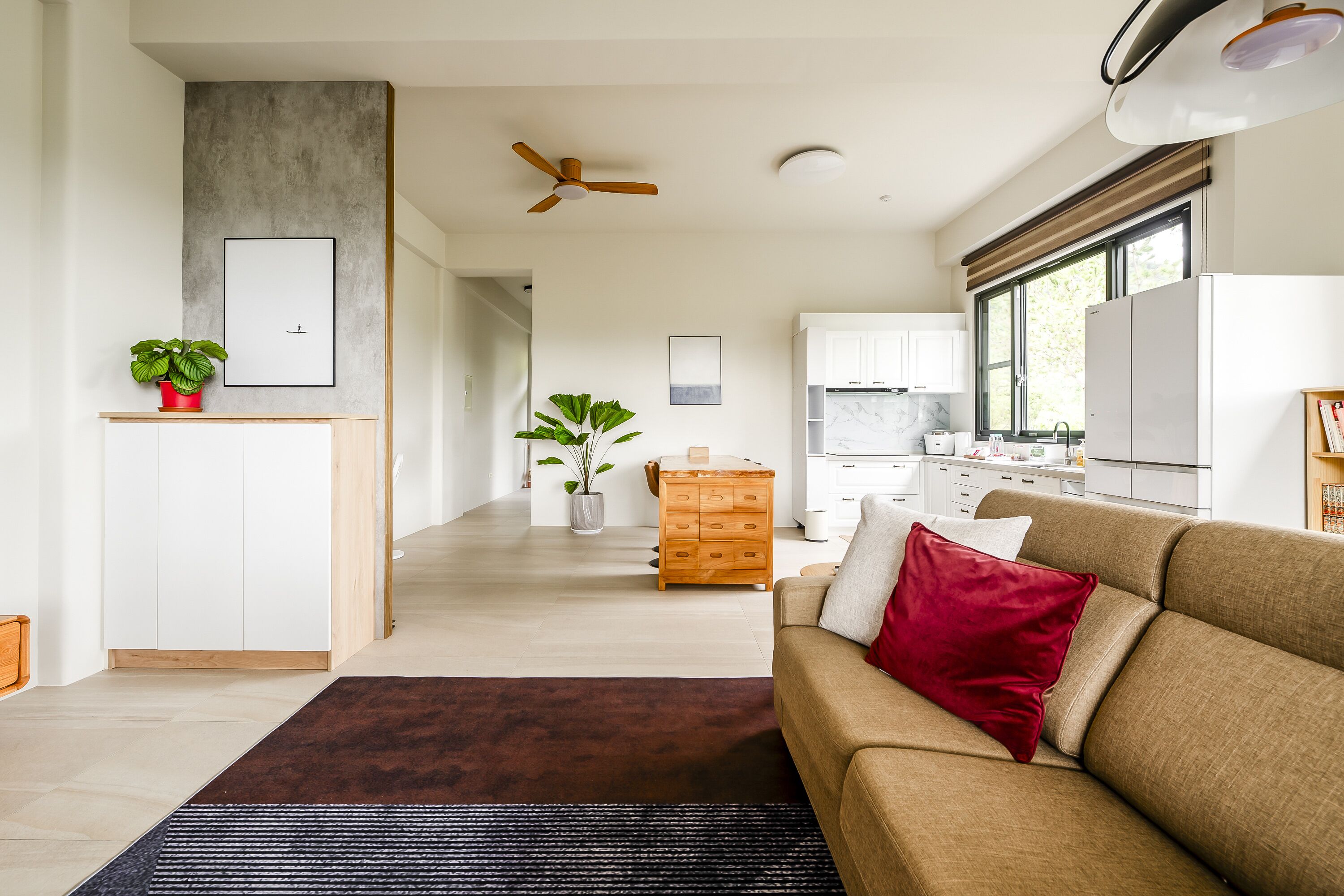 A modern living room with a sofa, a coffee table, and a red pillow. There is a wooden cabinet and a potted plant in the corner.