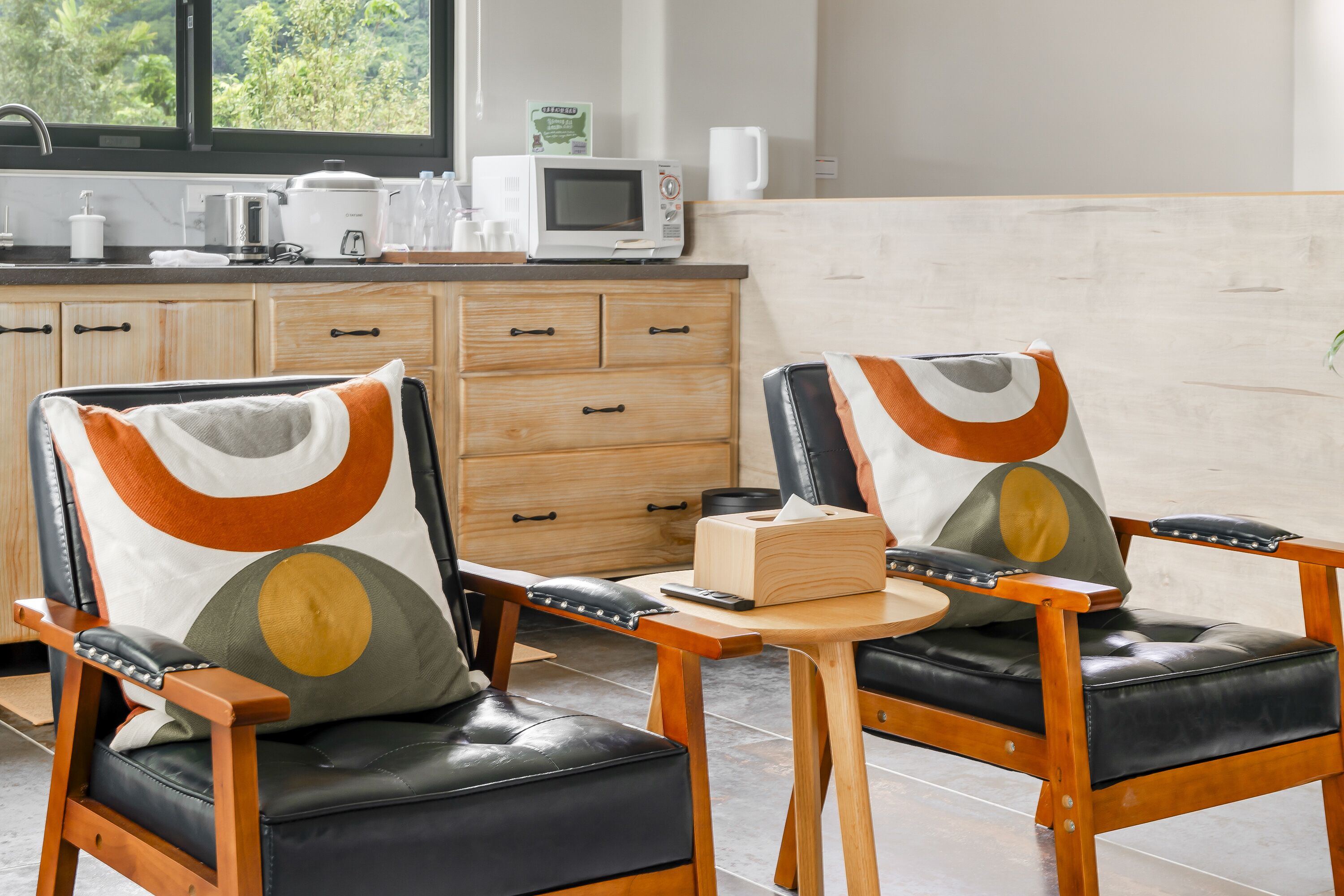 A modern kitchen with wooden cabinets, a microwave, and a coffee maker. There are two chairs with cushions and a small wooden table.