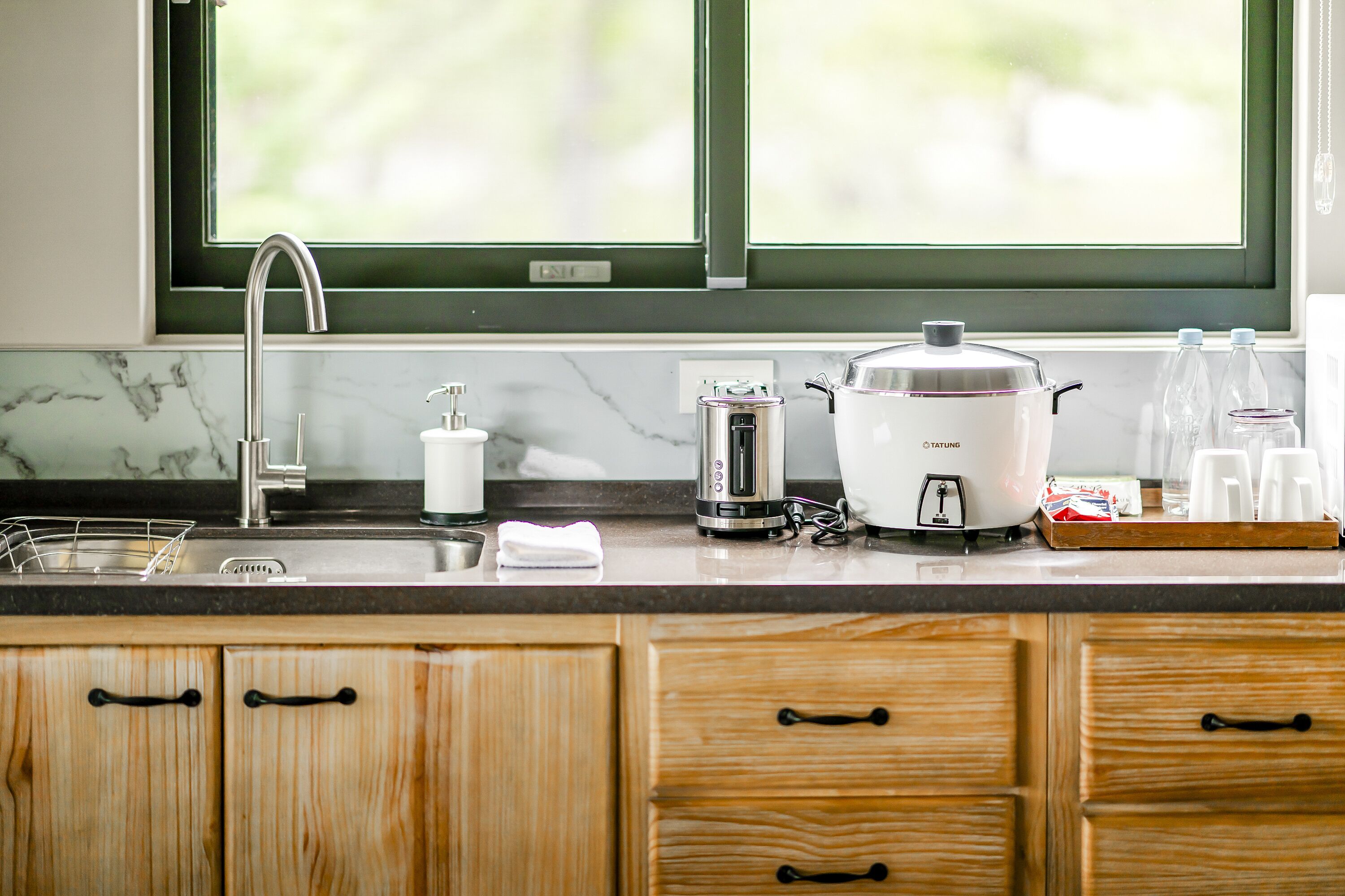 A kitchen with a sink, a coffee maker, a rice cooker, and a window with a view of greenery.