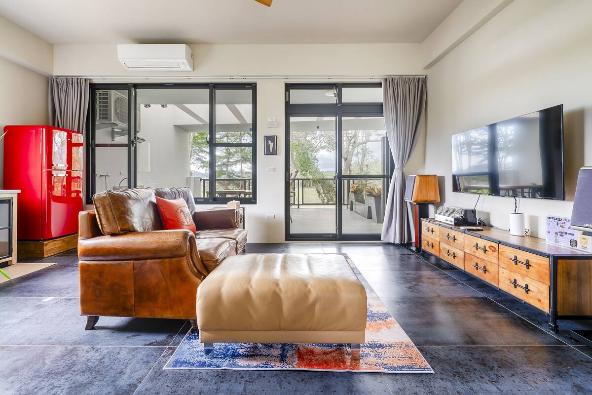 Uma sala de estar ampla com sofá de couro, um armário de madeira, uma televisão e uma grande porta de vidro deslizante que dá para uma área externa.
