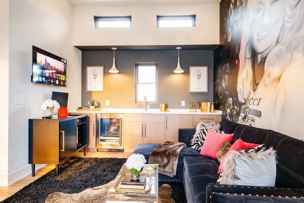 A modern living room with a black sofa, a coffee table with books and flowers, and a kitchen area with pendant lights and a TV.