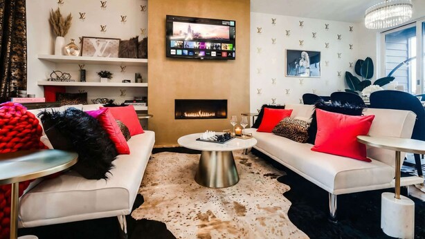 Un salon moderne avec une cheminée, des canapés blancs agrémentés de coussins rouges et noirs, un téléviseur fixé au mur et un lustre.