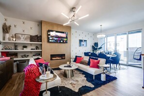 Un salon moderne avec une cheminée, un téléviseur fixé au mur et un coin repas avec des chaises bleues.