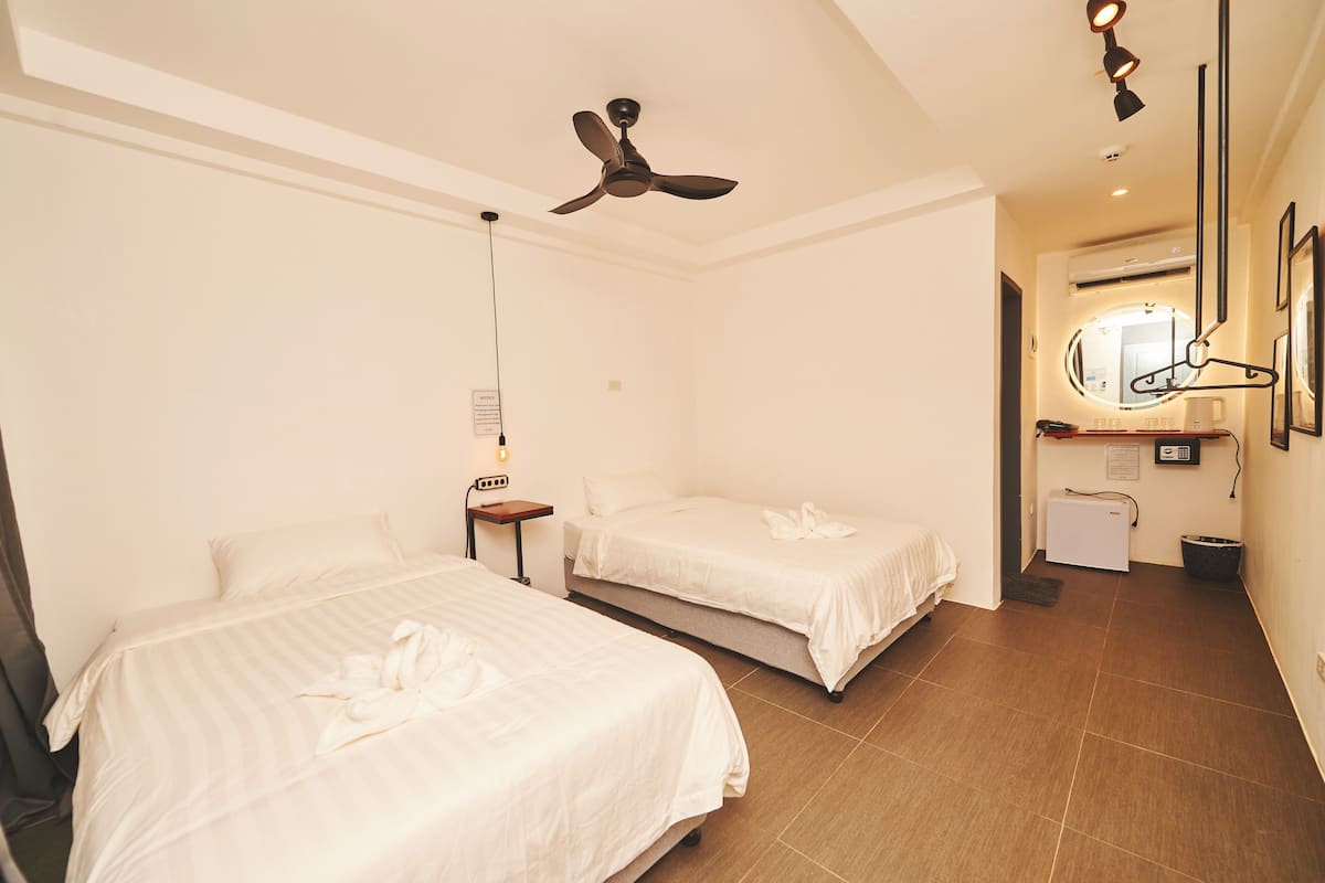 A hotel room with two beds, a ceiling fan, a small table with a lamp, a mirror, and a refrigerator.
