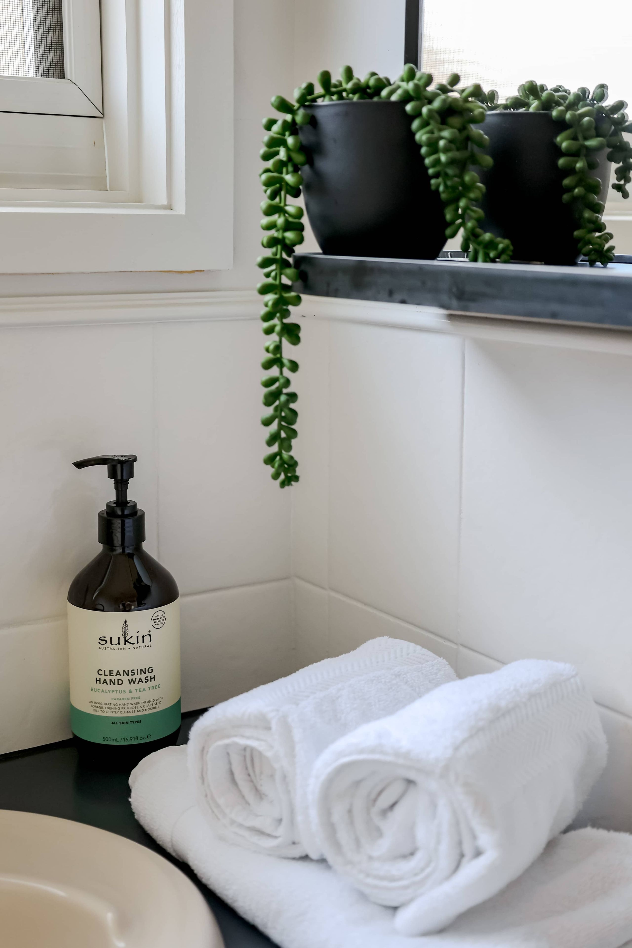 Une salle de bain avec un lavabo blanc, un flacon de savon liquide pour les mains de la marque SuKin, une serviette blanche roulée et trois plantes en pot disposées sur une étagère.
