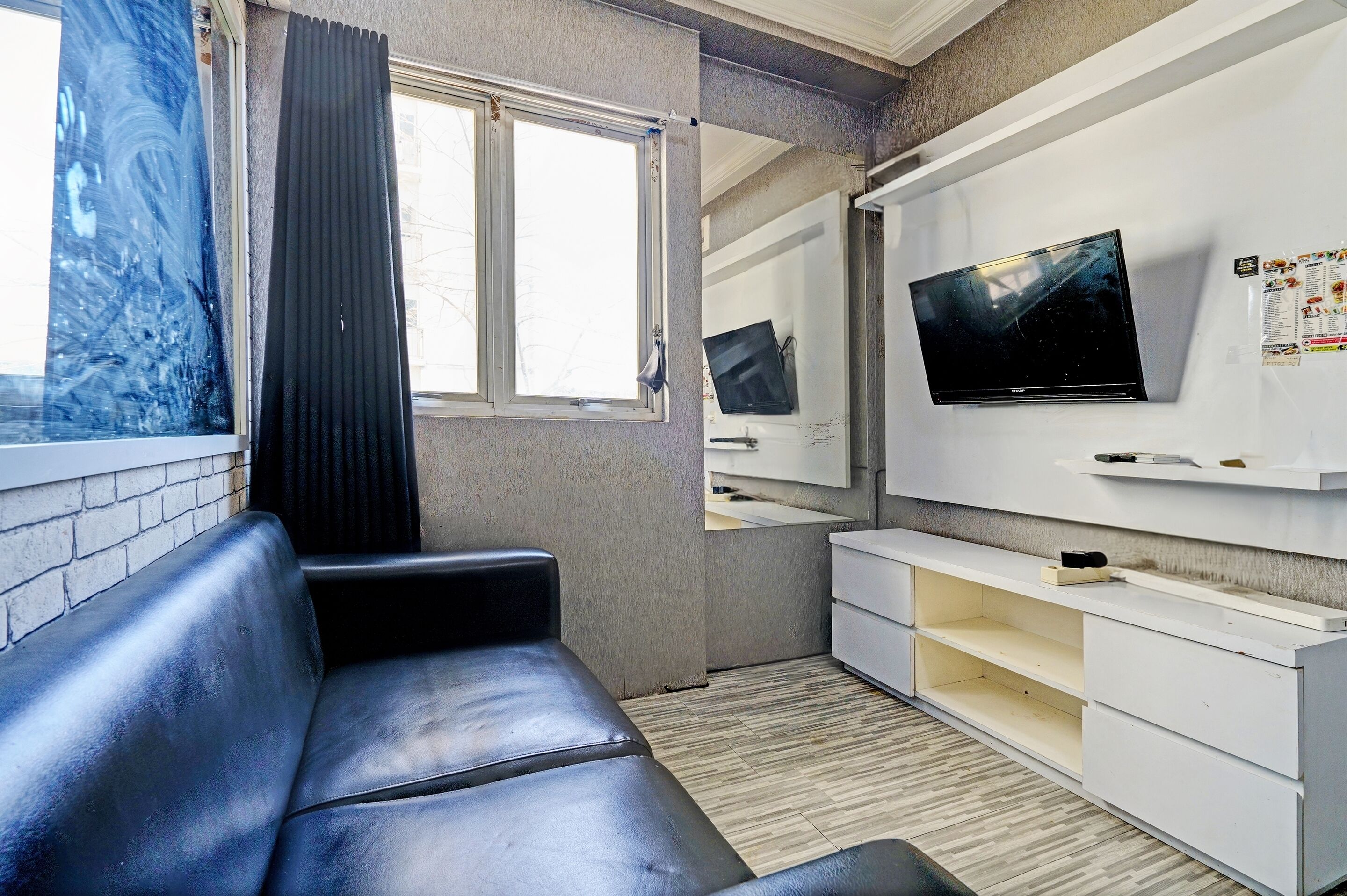 Een kleine, moderne kamer met een zwarte leren bank, een groot raam met uitzicht, een tv aan de muur en een wit kastje met spiegel.