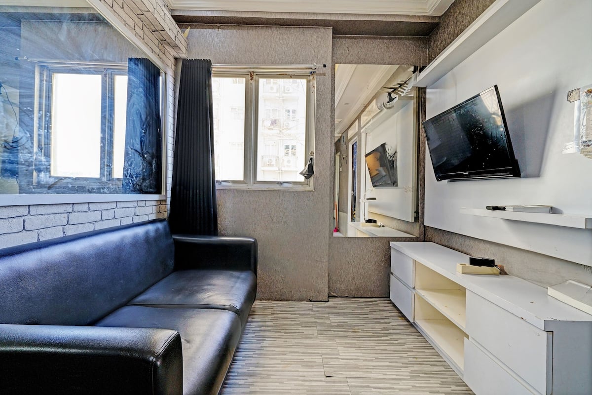 A modern living room with a black leather sofa, a flat-screen TV mounted on the wall, and a large window with curtains.