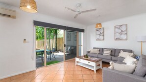 A modern living room with a grey sofa, a coffee table, and a ceiling fan. There is a sliding glass door leading to an outdoor area with a patio and a wooden fence. - 5 Reef Terraces Port Douglas (Port Douglas)