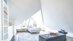 Un espacioso dormitorio en ático con un ventanal grande, una cama, un banco tapizado en gris y un estante blanco.