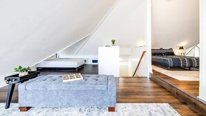 Un dormitorio estilo loft moderno con techo inclinado, un ventanal grande, una cama con ropa de cama estampada, un banco tapizado en azul y una mesita de noche blanca con una planta.
