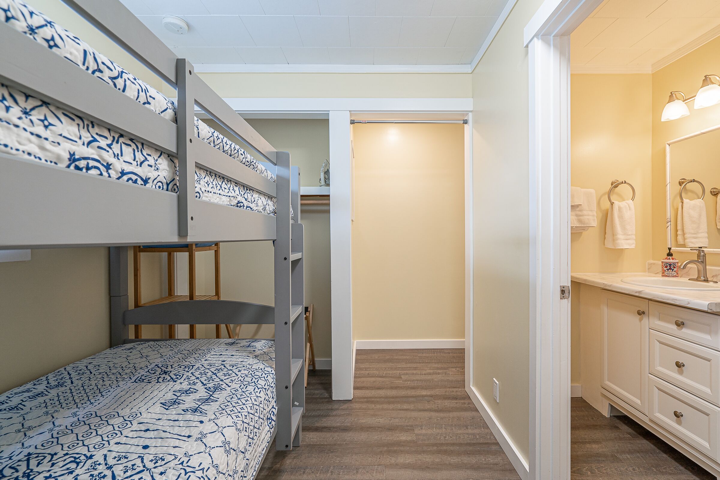 A bunk bed with patterned bedding, a bathroom with a sink and mirror, and a hallway leading to a closet.