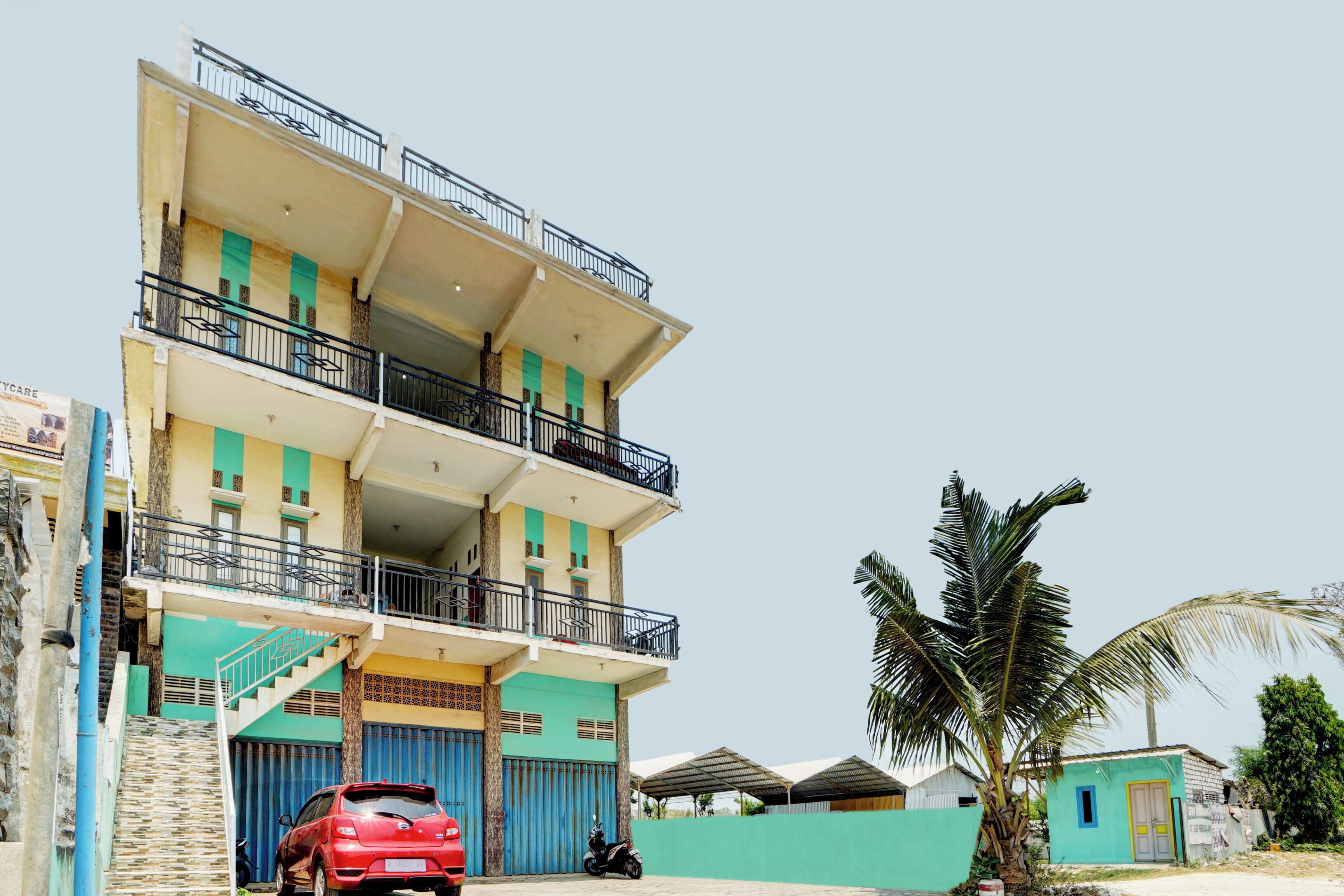 A multi-story building with a balcony, a red car parked in front, and a palm tree nearby.