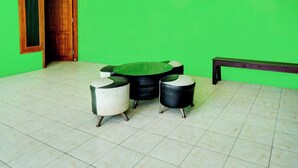 A room with a green wall, a wooden door, and a tiled floor. There is a round table with a black top and two chairs with white and black seats. A bench is against the wall. - OYO Life 93075 Nova Jaya 2 Homestay Syariah (Paciran)