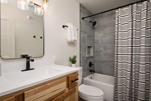 Un bagno moderno con un lavandino bianco, un mobile contenitore in legno, un WC bianco, un box doccia in vetro e una tenda da doccia a righe.