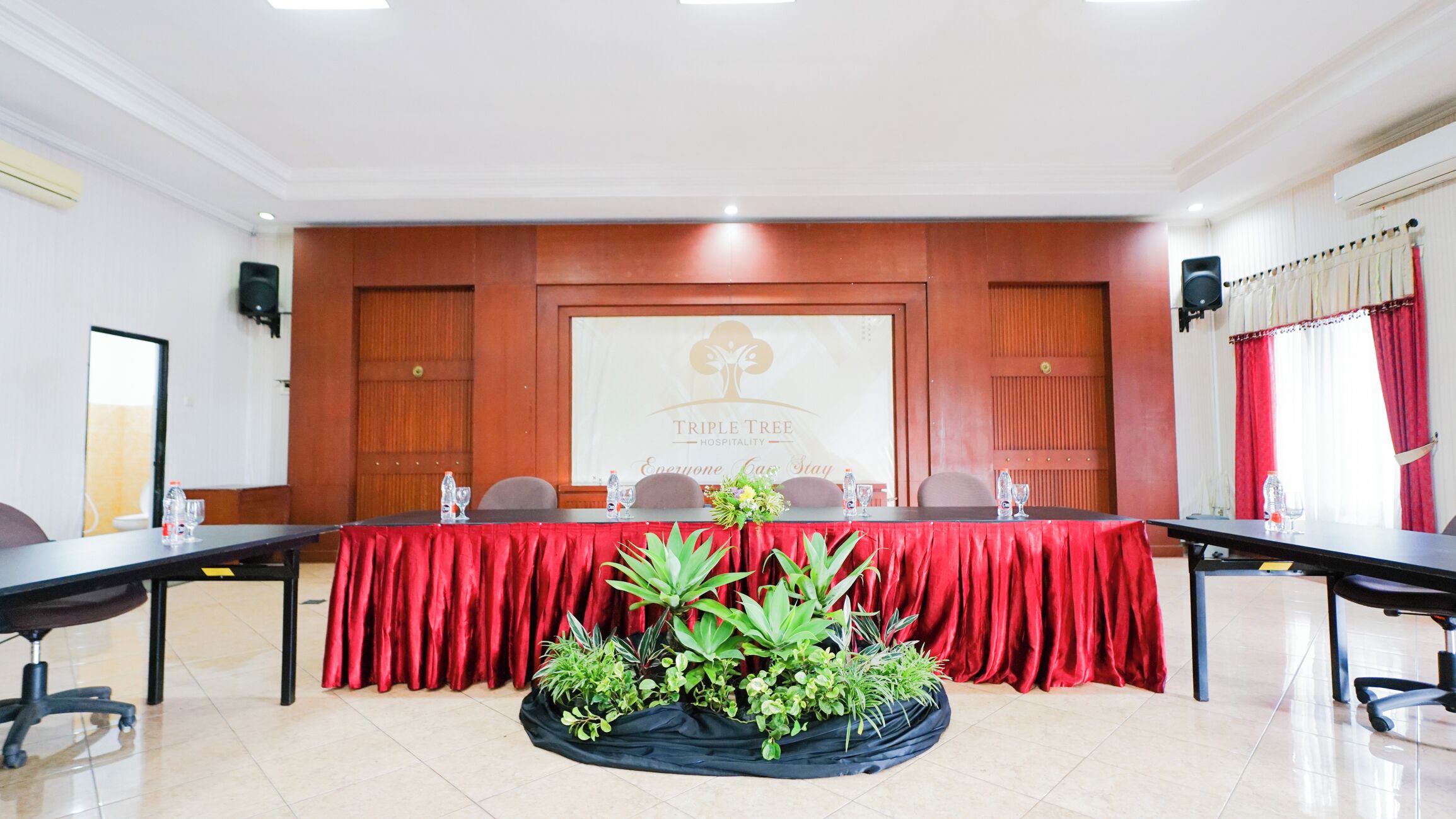 Un salón de conferencias con una larga mesa, sillas, un mantel rojo, plantas en macetas y un cartel con los textos 'TRIPLE TREE' y 'Everyone Care Stay'.