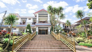 A multi-story building with a central entrance, surrounded by a garden with palm trees and other plants. - Tripletree Sinergi Hotel Tretes (Prigen)
