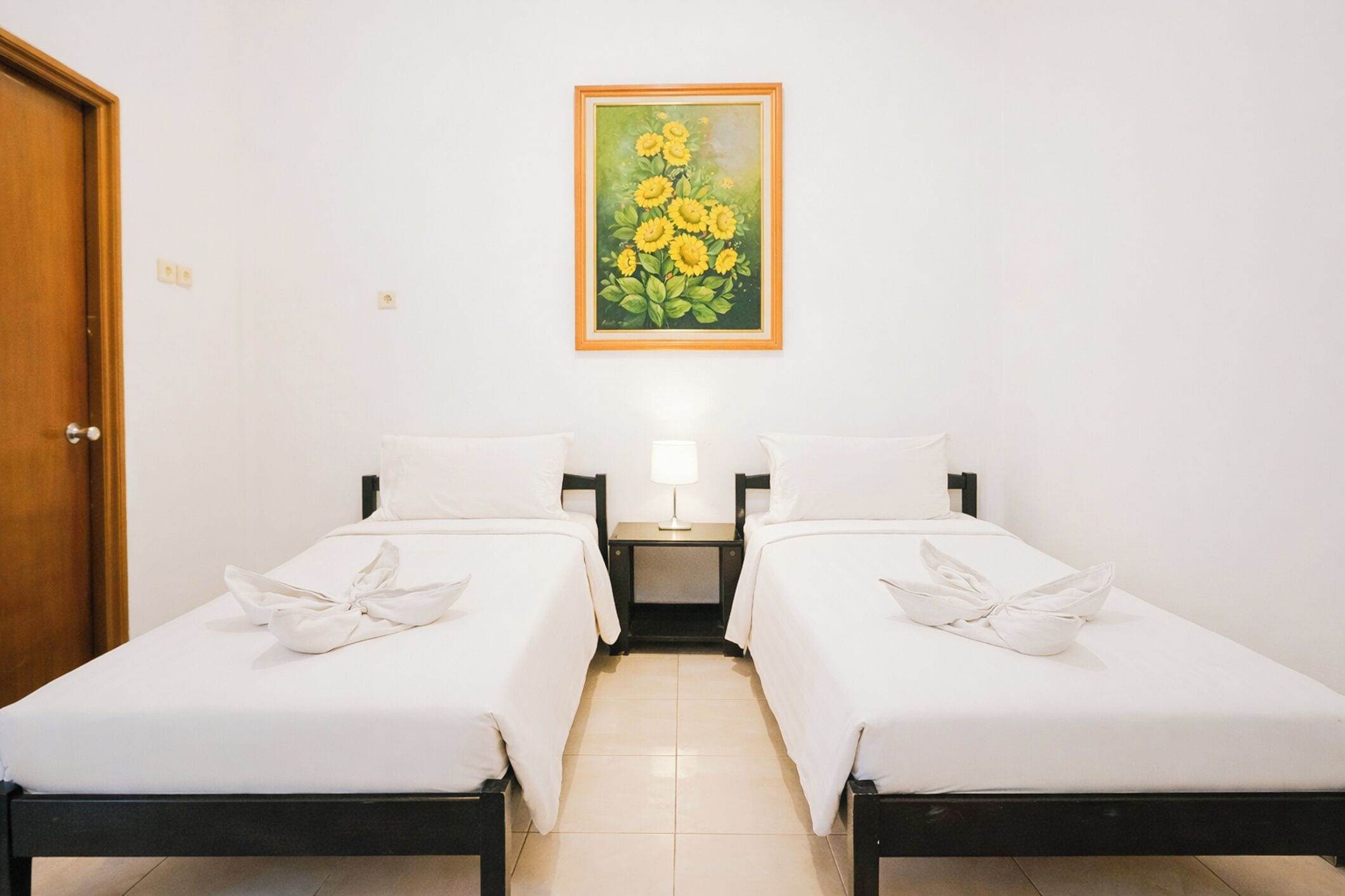 Habitación de hotel con dos camas, una mesita de noche con lámpara y un cuadro de flores en la pared.