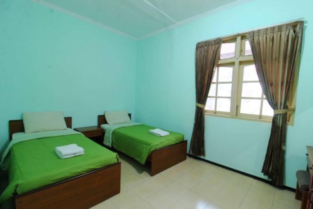 A room with two beds, each with green bedding and white pillows, a window with curtains, and a tiled floor.