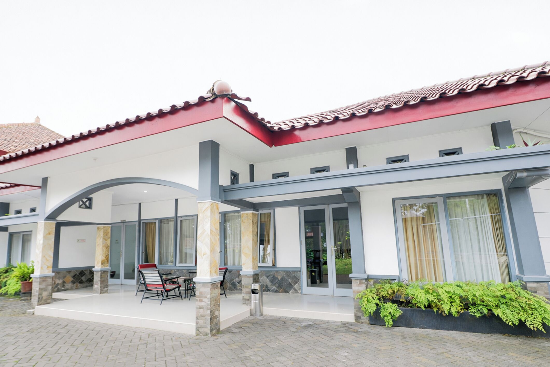 A hotel with a red-tiled roof, a covered outdoor seating area with a table and chairs, and a landscaped garden.