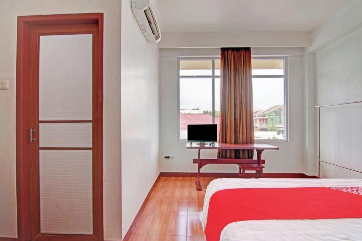 Ein Hotelzimmer mit einem Bett, einem Schreibtisch mit Fernseher, einem Fenster mit Vorhängen und einer Klimaanlage.