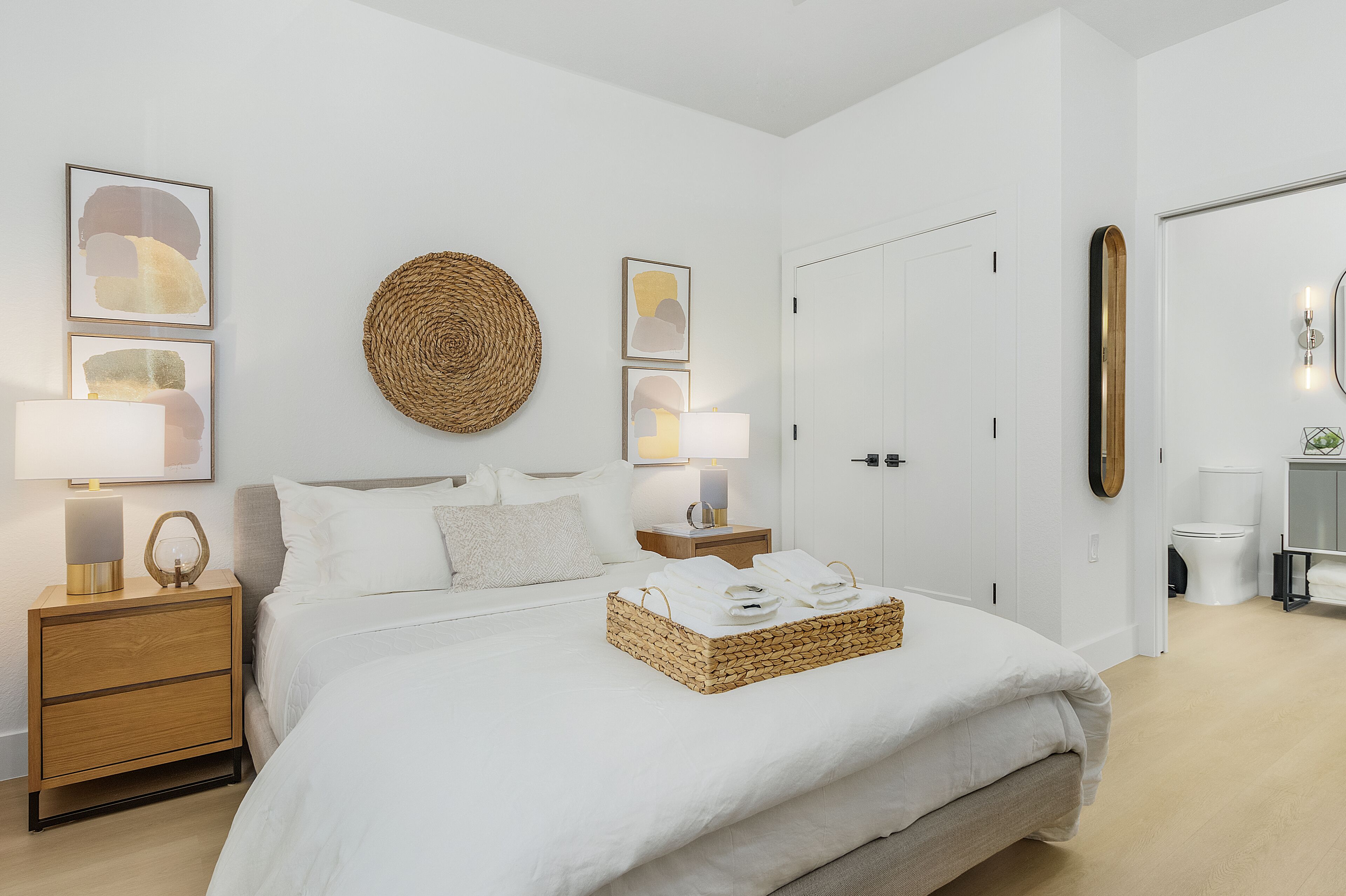 A modern bedroom with a large bed, a bedside table with a lamp, and a wall-mounted round mirror.