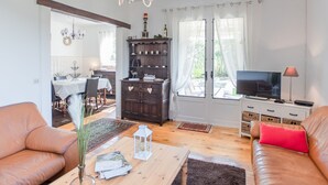 Een woonkamer met een bank, een salontafel, een televisie en een houten kast. Achtergrond toont een eethoek met een tafel en stoelen.
