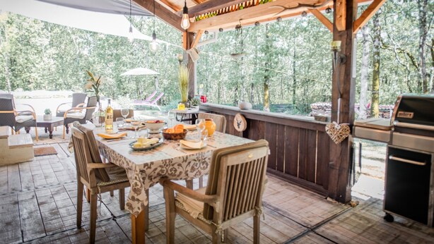 Een terras met houten tafels en stoelen, een barbecue en uitzicht op bomen.