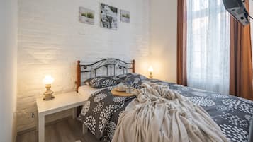 Uma cama bem arrumada com um cobre-leito estampado, duas luminárias de cabeceira e uma mesinha com xícara e pires.