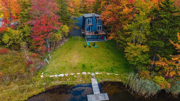 Ein zweigeschossiges Haus mit einer Terrasse und einem Steg, der sich über das Wasser erstreckt, umgeben von Bäumen im Herbstlaub.