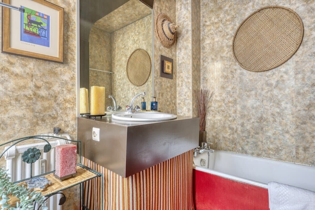 A bathroom with a marble wall, a large mirror, a sink with two faucets, a red bathtub, and striped towels.