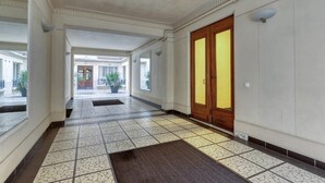 Um corredor com piso de azulejos, porta de madeira com painéis de vidro e vista de um edifício ao fundo.