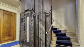 Uma escadaria imponente com guarda-corpo de metal e degraus cobertos por carpete azul, que levam a um patamar.