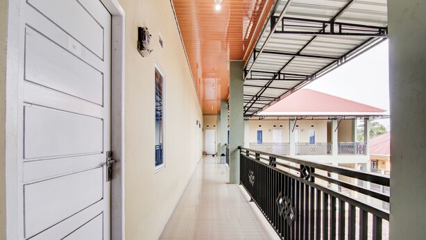 A corridor with a white door, a balcony with a metal railing, and a red-roofed building in the background. - OYO 93087 Wisma Apel Syariah (Pekanbaru)