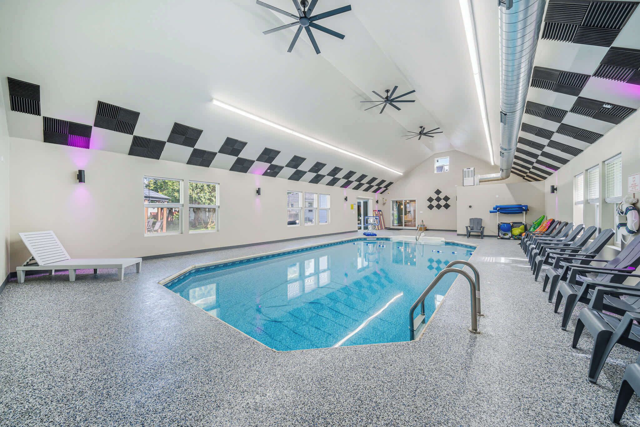 A modern indoor swimming pool with a clear blue water, surrounded by lounge chairs and a ceiling with geometric patterns and ceiling fans.